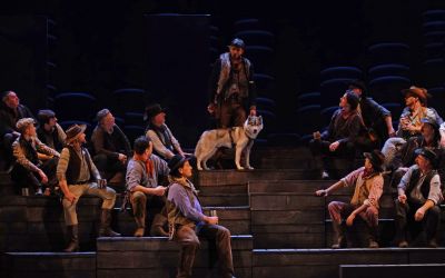 na drewnianych uniesieniach stoi mężczyzna z dużym psem, wokół niego siedzą mężczyźni którzy mu się przypatrują na ciemnym tle widać zarysy dwóch prostokątów oraz kilkunastu dysków