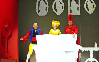 na pierwszym planie biała kanapa za którą stoją trzy osoby, mężczyzna ubrany na kolorowo śpiewa, kobieta ubrana na żółto oraz mężczyzna ubrany na czerwono mają zdziwione miny, w tle białe obrazy oraz czerwona klatka dla ptaka, szare tło