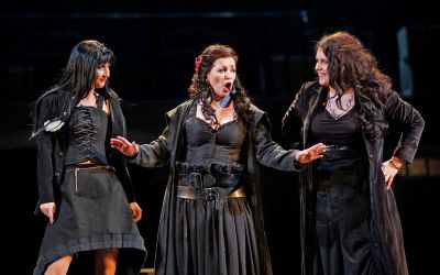 trzy kobiety ubrane na czarno, stojąca na środku śpiewa, a dwie pozostałe wpatrują się w siebie szeroko uśmiechnięte, czarne tło