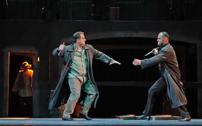 na scenie stoi dwóch mężczyzn ubranych na szaro z nożami w rękach, walczą ze sobą, w tle widać przejście w którym stoi inny mężczyzna który przypatruje się całej sytuacji, ciemne tło