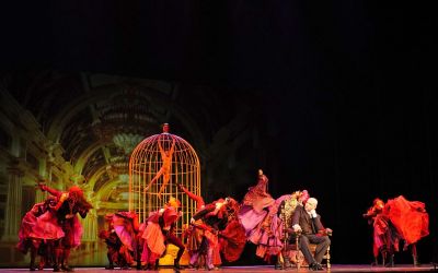 scena grupowa, tancerze ubrani w czerwone stroje tańczą, po ich prawej złoty fotel, na którym siedzi mężczyzna w garniturze i się im przypatruje, w tle żółta klatka w której tańczy ubrany na żółto tancerz, czarne tło