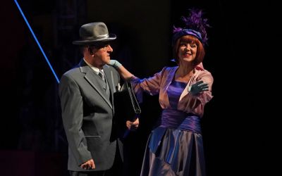 czarne tło, po lewej mężczyzna ubrany w szary garnitur, kapelusz na głowie w lewej ręce pod pachą dzierży skórzaną teczkę patrzy na kobietę, kobieta stoi po prawej i trzyma mężczyznę za ramię, jest ubrana w odcieniach różu i fioletu, ma błękitne rękawiczki, fioletowe zdobione nakrycie głowy, rude włosy i czerwoną szminkę, jest uśmiechnięta