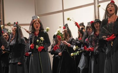 grupa kobiet w szarych strojach i welonach, mają czerwone rękawiczki, trzymają białe róże w rękach i śpiewają, w tle szyby
