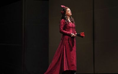 kobieta w długiej bordowej sukni patrzy skupiona przed siebie, odwrócona w prawą stronę, w rękach trzyma czarno-czerwony, złożony wachlarz, ciemne tło