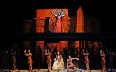scena grupowa na dole sceny stoi około czterdziestu aktorów, na pierwszym planie mężczyzna ubrany w białą szatę, złotą pelerynę oraz złoty, wielki naszyjnik, ma intensywnie pomalowane oczy na czarno, po jego prawej i lewej stronie po trzy tańczące kobiety w krótkich, brązowych sukienkach z czerwonymi zdobieniami, reszta aktorów w tle, nad sceną stoi śpiewająca solistka ubrana w czarną długą, wąską sukienkę, ma także ogon pawia i dużo biżuterii - na głowie oraz szyi