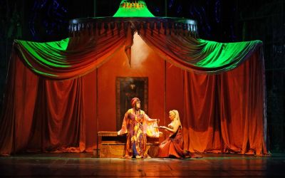 duży pomarańczowy namiot od góry oświetlony zielonym światłem, w środku stół na którym siedzi mężczyzna ubrany w fioletowo-złotą szatę, ma czerwony turban na głowie, po prawej kobieta klęczy przed mężczyzną i przekazuje mu płaski przedmiot, jest ubrana w brązową sukienkę zdobioną biżuterią na głowie ma opaskę, w tle namiotu wisi ciemny obraz z niewyraźną postacią