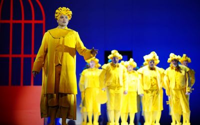 z przodu po lewej śpiewający solista, w z tyłu po prawej grupa stojących osób, wszyscy ubrani w intensywnie żółte stroje, tło jest niebieskie