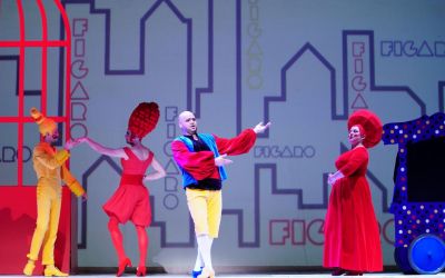 w tle miasto, z losowymi napisami figaro w różnych miejscach, a na scenie czwórka aktorów, z lewej tańcząca para ubrana na żółto oraz czerwono, po prawej ubrana na czerwono kobieta wpatrująca się w solistę ubranego na kolorowo, który śpiewa