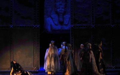 w tle żelazna spawana ściana na jej środku szeroka dziura przez którą przechodzi grupa aktorów ubrana na kremowo, nad nimi wisi dekoracyjna wyrzeźbiona w głowa faraona, po lewej stronie od przechodzących aktorów siedzi na kolanach ubolewająca kobieta, podpiera się ręką sceny i patrzy w górę z wyrazem bólu na twarzy, oświetlenie sceny jest niebieskie