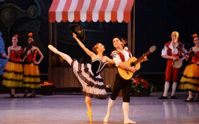 na pierwszym planie para, mężczyzna z gra na gitarze kobieta uśmiechnięta tańczy opierając się o niego, w tle stragan z owocami, po jego lewej i prawej stronie aktorzy ubrani w ciepłe kolory w hiszpańskim stylu
