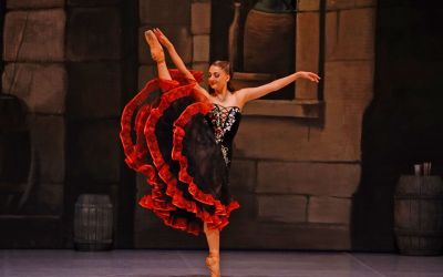 tańcząca kobieta unosząca nogę bardzo wysoko, ubrana w czarną suknię z czerwonymi zdobieniami, w tle ściana z kamiennych bloków, beczki