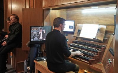organista gra, po jego lewej monitor na którym widać Rafała Janiaka, w tle siedzi dwóch mężczyzn ubranych na biało, wokół drewniane ściany