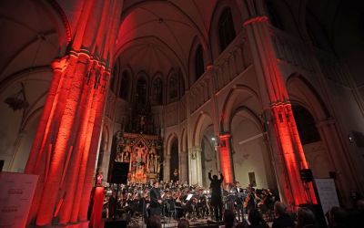 grupa chórzystów i orkiestra z dyrygentem z perspektywy widowni w kościele po lewej i prawej oświetlone na czerwono filary