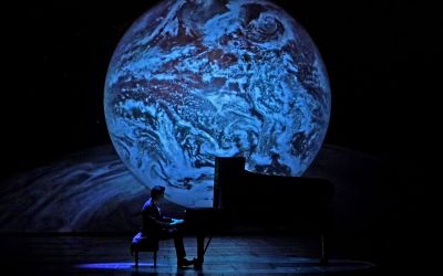 mężczyzna siedzi przy fortepianie i na nim gra, w tle niebieska planeta na czarnym tle
