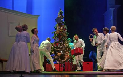 na środku świątecznie przyozdobiona choinka, pod nią czerwone prezenty wokół aktorzy ubrani na biało