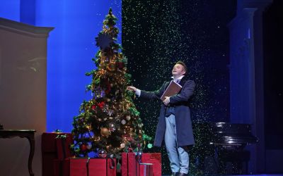 solista w czarnym płaszczu z brązową teczką w ręce stoi po prawej stronie od świątecznie przyozdobionej choinki z czerwonymi prezentami pod nią i śpiewa, w góry leci na niego śnieg, tło jest niebieskie