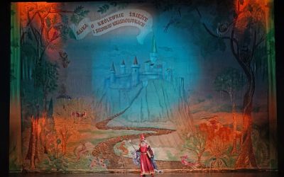 czerwony czarodziej stoi na środku sceny z berłem, w tle drzewa, ścieżka do zamku na wzgórzu i napis u góry 