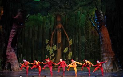 siedmiu krasnoludków w czerwonych strojach trzymają się za ręce i tańczą, w tyle trzy drzewa z uśmiechami, w tle las