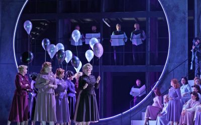 Na pierwszym planie kobiety trzymające w dłoniach fioletowo - białe balony. Kobiety są ubrane w długie spódnice, mają spięte włosy. Po prawej stronie kobiety siedzą na schodach. W tle mężczyźni stojący na balkonie, w dłoniach trzymają rozłożone gazety. 