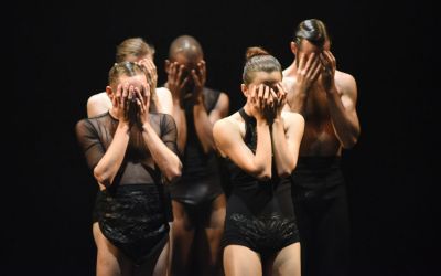 grupa tancerzy w czarnych strojach na czarnym tle zasłaniająca sobie twarze dłońmi