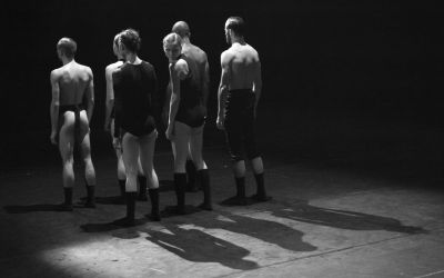 siódemka tancerzy stoi tyłem w czarnym stroju, jedna tancerka obraca głowę, czarne tło