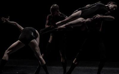 czwórka tancerzy w różnych pozach, dwóch przytrzymuje kobietę w powietrzu, jeden po lewej jest odchylony do tyłu, czarne tło, czarne stroje