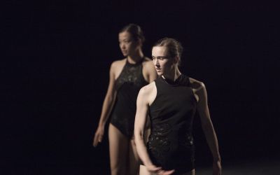 dwie tancerki w czarnych strojach na czarnym tle stoją prosto, mają głowy odwrócone w lewą stronę