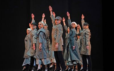 Scena zbiorowa. Grupa tancerzy ubrana w szare płaszcze, nakrycia głowy. Wszyscy mają uniesione wysoko dłonie. 