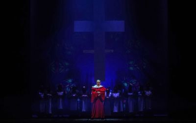 Starszy mężczyzna w czerwonej szacie na tle wysokiego krzyża. Za jego plecami ubrani na biało chórzyści. 