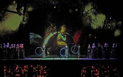 Na środku sceny 3 aktorów z obręczami w dłoniach. Czarna scenografia z elementami gałęzi drzew i jasnym płomieniem. Z dwóch stron chórzyści w czarnych kostiumach. 