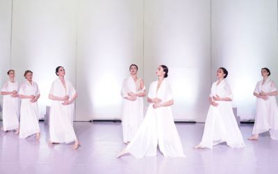 siedem kobiet w białych sukniach patrzy w górę i śpiewa na tle białej ściany