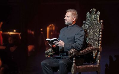 Mężczyzna z brodą siedzi na krześle. W dłoniach trzyma otwartą książkę. Ubrany jest w czarną koszulę i ciemne spodnie. Jest zamyślony i skupiony. 
