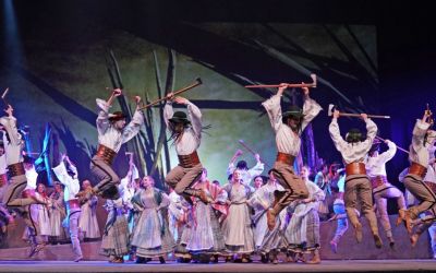 na pierwszym planie skaczący górale z ciupagami, za nimi duża grupa tańczących kobiet ubranych w jasne suknie, w tle scenografia z drewnianymi palami, niebieskie oświetlenie