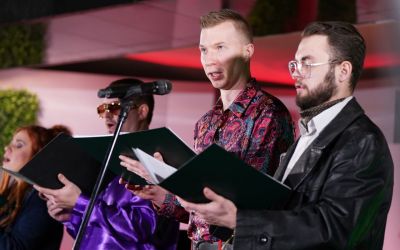 Na zdjęciu śpiewający mężczyźni. Z prawej strony artysta w okularach i skórzanej kurtce, kolejny mężczyzna we wzorzystej koszuli, ostatni w okularach przeciwsłonecznych. 
