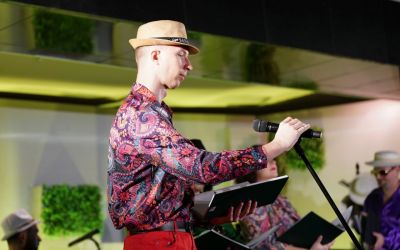 Na zdjęciu mężczyzna trzymający mikrofon na statywie. Jest ubrany we wzorzystą koszulę, czerwone spodnie i kapelusz na głowie. W tle scenografia z motywem zieleni. 