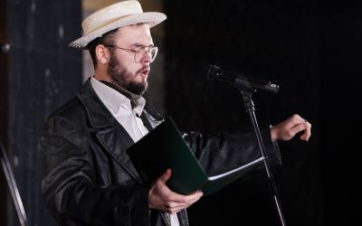 Na zdjęciu mężczyzna śpiewający do mikrofonu. Ubrany w czarną skórzaną kurtkę i białą koszulę, na głowie ma słomkowy kapelusz. 