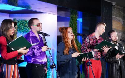 Scena zbiorowa. Od prawej: kobieta w czerwonej bluzce, spódnicy w paski, dalej mężczyzna w fioletowej koszuli, okularach przeciwsłonecznych, kobieta w rudych włosach, mężczyzna w czerwonych spodniach i wzorzystej koszuli, na końcu mężczyzna w skórzanej kurtce. 