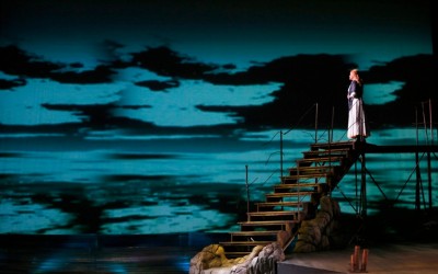 na schodach stoi kobieta, wpatruje się w niebieskie niebo oraz wodę w tle, na dole zdjęcia drewniany pomost