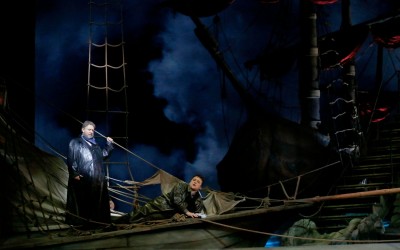 na łodzi stoi mężczyzna trzymając jedną z lin, drugi mężczyzna leży pod nim i śpiewa, z tyłu stoi druga łódź, w tle chmury, dużo lin i żagli