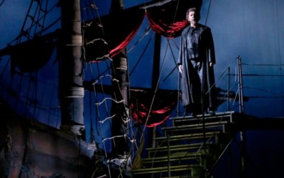 mężczyzna stoi na schodach nieopodal dużego statku, ubrany w długi czarny płaszcz wpatruje się w przestrzeń, niebieskie tło, dużo lin i żagli