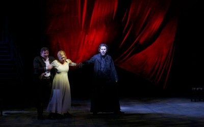 trójka aktorów, dwóch mężczyzn ubranych na czarno, między nimi ubrana na biało, kulejąca kobieta, z bólem na twarzy, w tle czerwona kurtyna