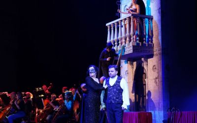 po lewej orkiestra, po prawej ściana z balkonem na którym stoi kobieta, pod balkonem drabina po której wspina się mężczyzna w czerni, na pierwszym planie para ubrana w odświętne stroje i patrząca w prawą stronę, czarne tło