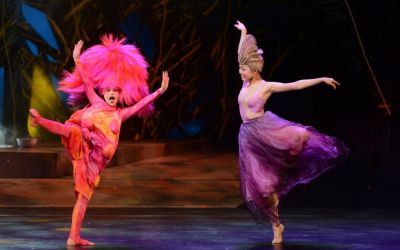 dwie tancerki unoszące wysoko nogi oraz ręce, jedna ubrana w różowy strój z dużą peruką, druga ubrana w fioletową suknię, w tle wodorosty i drewniane podwyższenie 