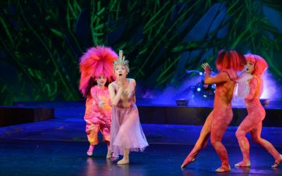 z przodu tancerka w fioletowej sukience, za nią trzy tancerki ubrane w różowe stroje i peruki, tańczą i mają zdziwione miny, w tle wodorosty, oświetlenie niebieskie